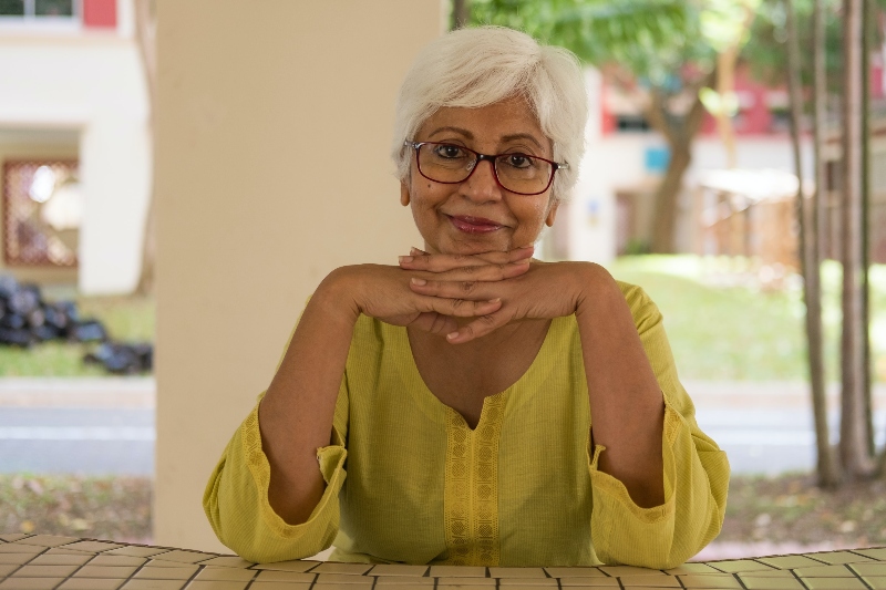 A senior woman is posing for the picture sitting at a table outside with her hands under her chin smiling.