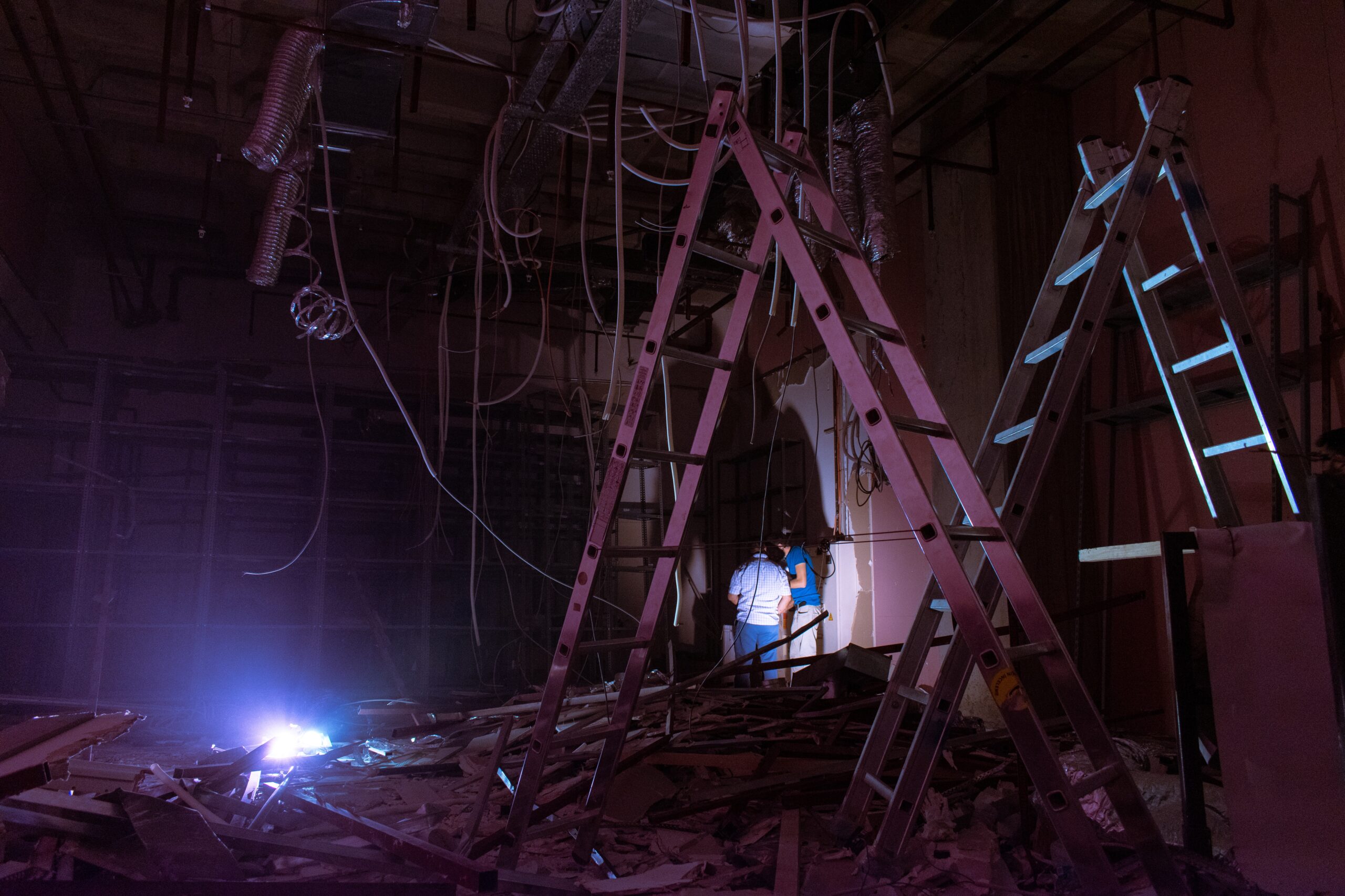 A flood, fire or disaster happened. There are wires, cords and pipes handing from the ceiling, debris all over the floor. There is no power, two people are looking around with flashlights